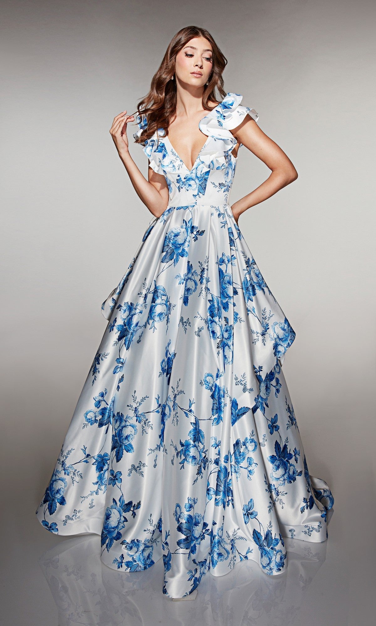 A woman poses against a plain light background wearing the Alyce 62001 long prom dress in blue/white, featuring blue floral prints, ruffled sleeves, and a deep V-neckline.